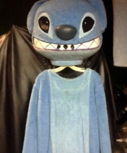 Blue furry costume with large head and big eyes on a hanger against a dark background.