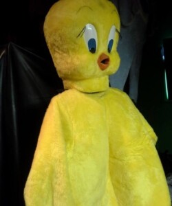 Person wearing a large yellow bird costume with oversized eyes and an orange beak, standing against a dark background.